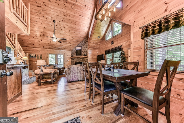 dining space featuring a stone fireplace, light hardwood / wood-style floors, wooden walls, high vaulted ceiling, and ceiling fan