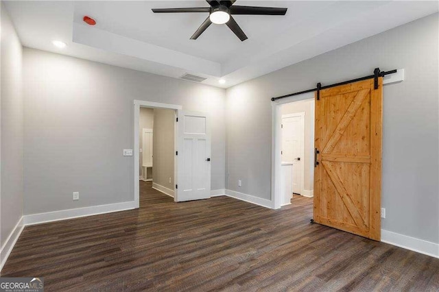 empty room with a tray ceiling, a barn door, ceiling fan, and dark hardwood / wood-style floors