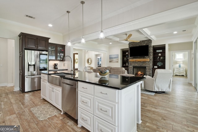 kitchen with a kitchen island with sink, stainless steel appliances, a stone fireplace, white cabinetry, and light hardwood / wood-style flooring