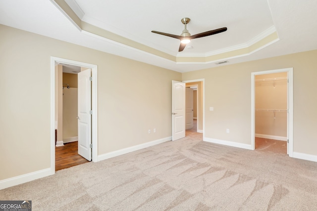 unfurnished bedroom with ceiling fan, a spacious closet, a raised ceiling, crown molding, and light colored carpet