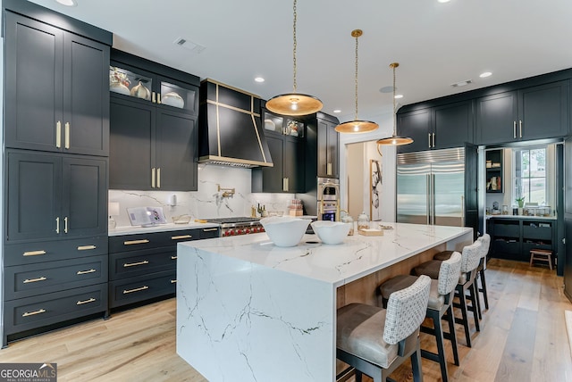 kitchen featuring a large island with sink, wall chimney exhaust hood, stainless steel built in refrigerator, hanging light fixtures, and light hardwood / wood-style flooring