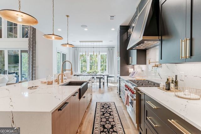 kitchen featuring a wealth of natural light, a kitchen island with sink, stainless steel range, wall chimney range hood, and decorative light fixtures