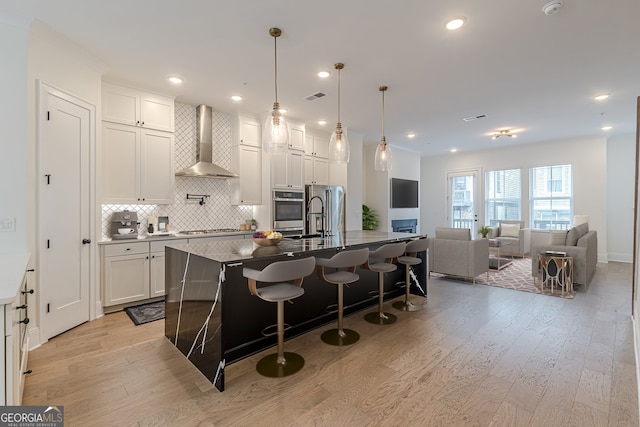 kitchen featuring light hardwood / wood-style flooring, a kitchen breakfast bar, wall chimney range hood, appliances with stainless steel finishes, and a large island with sink