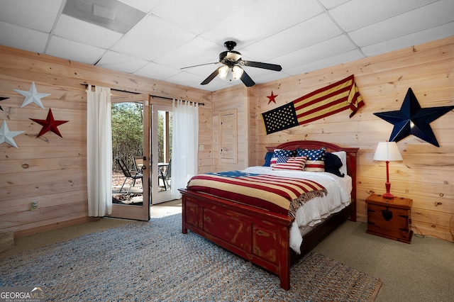 carpeted bedroom featuring access to outside, a paneled ceiling, ceiling fan, and wooden walls