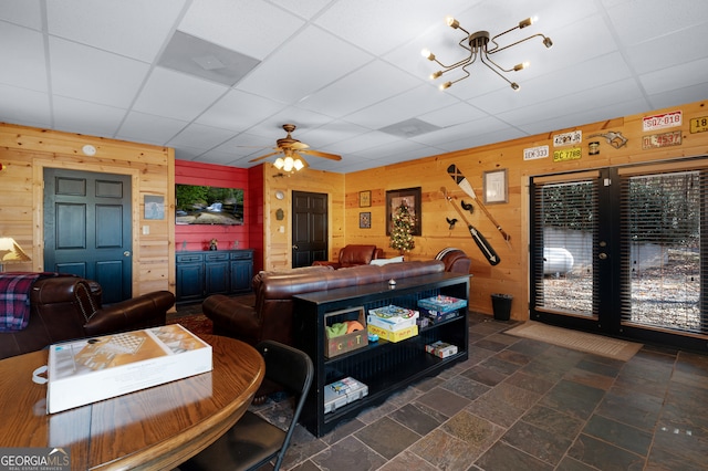 living room with a paneled ceiling, wood walls, and ceiling fan with notable chandelier