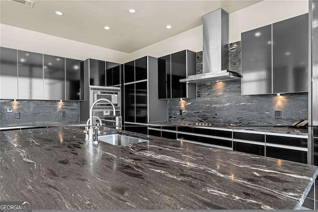 kitchen featuring sink, tasteful backsplash, dark stone countertops, and wall chimney range hood