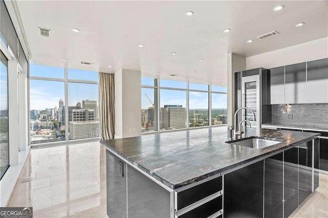 kitchen with tasteful backsplash, sink, a large island with sink, a wall of windows, and dark stone countertops