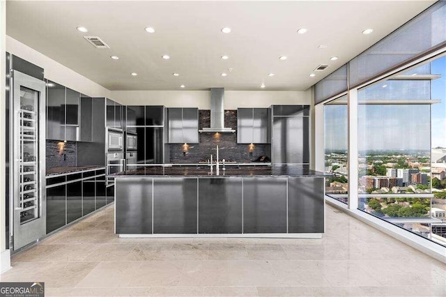 kitchen featuring backsplash, an island with sink, wall chimney range hood, and a wall of windows