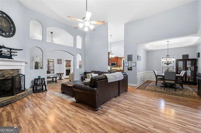 living room with a high ceiling, a fireplace, light wood-type flooring, and ceiling fan with notable chandelier