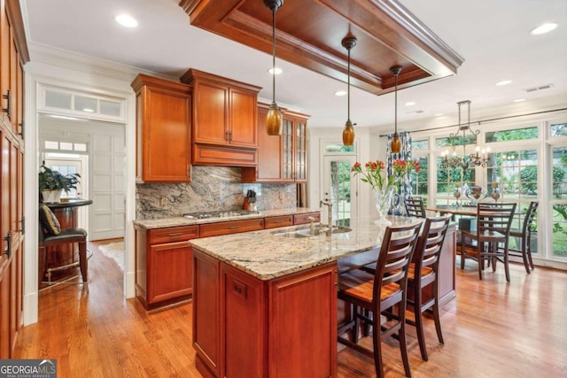 kitchen featuring pendant lighting, light stone countertops, light hardwood / wood-style floors, a tray ceiling, and a kitchen island with sink