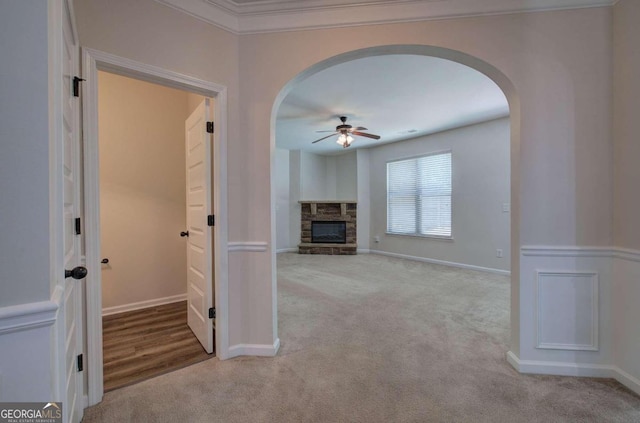 unfurnished living room featuring ceiling fan, a stone fireplace, light carpet, and ornamental molding