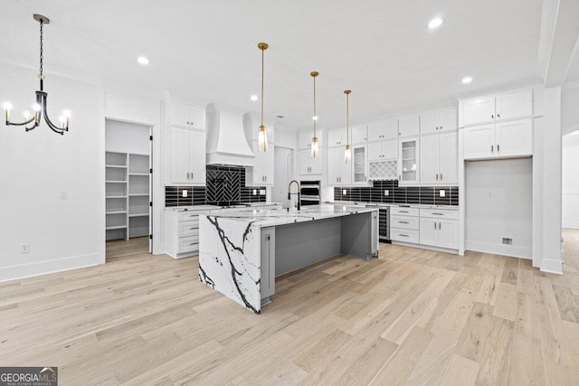 kitchen with light hardwood / wood-style floors, light stone counters, a kitchen island with sink, white cabinets, and premium range hood