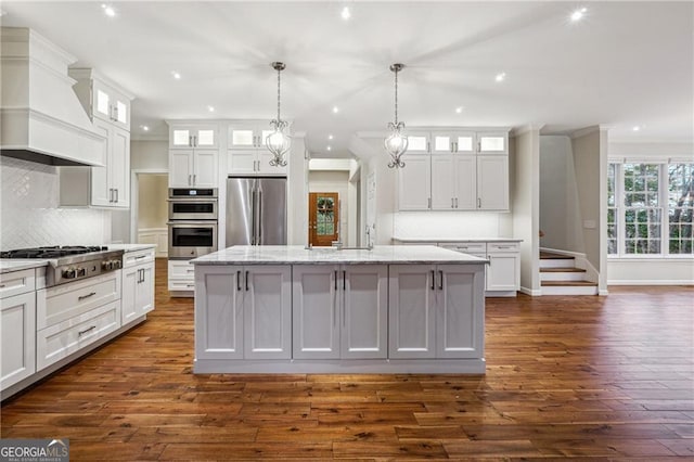 kitchen featuring custom exhaust hood, white cabinets, an island with sink, and stainless steel appliances