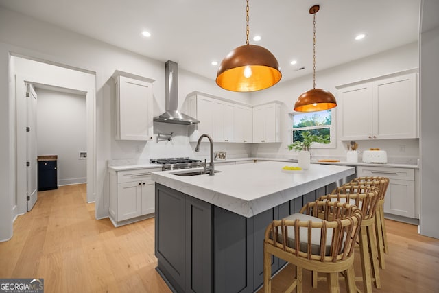 kitchen with a center island with sink, wall chimney range hood, decorative light fixtures, light hardwood / wood-style floors, and white cabinets