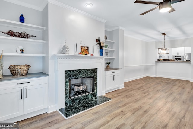 living room featuring light hardwood / wood-style floors, built in shelves, a premium fireplace, ceiling fan, and crown molding