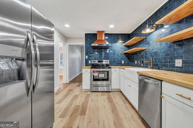 kitchen with white cabinets, wall chimney exhaust hood, light wood-type flooring, butcher block counters, and stainless steel appliances