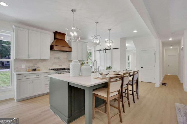 kitchen with premium range hood, a center island with sink, decorative light fixtures, white cabinets, and light hardwood / wood-style flooring