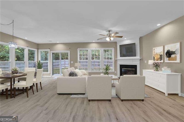 living room with a large fireplace, ceiling fan with notable chandelier, a healthy amount of sunlight, and light wood-type flooring