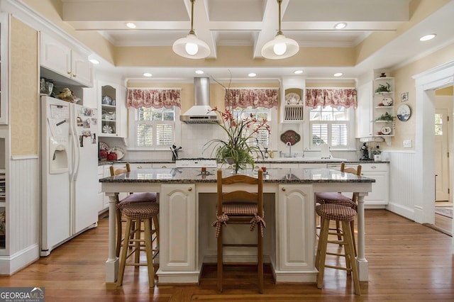 kitchen with dark stone counters, white refrigerator with ice dispenser, wall chimney exhaust hood, and open shelves