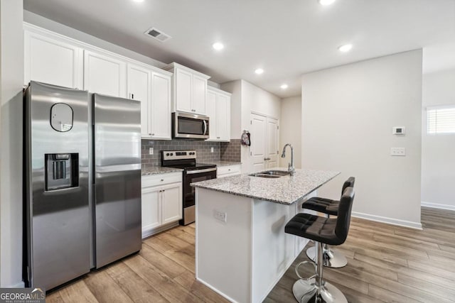 kitchen featuring light stone counters, a center island with sink, appliances with stainless steel finishes, white cabinets, and light hardwood / wood-style flooring