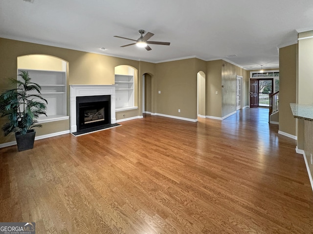 unfurnished living room featuring wood-type flooring, built in features, ceiling fan, and crown molding