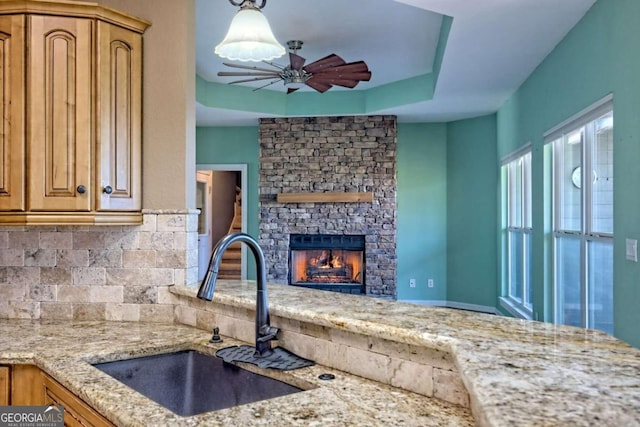 kitchen featuring sink, light stone countertops, ceiling fan, backsplash, and a stone fireplace