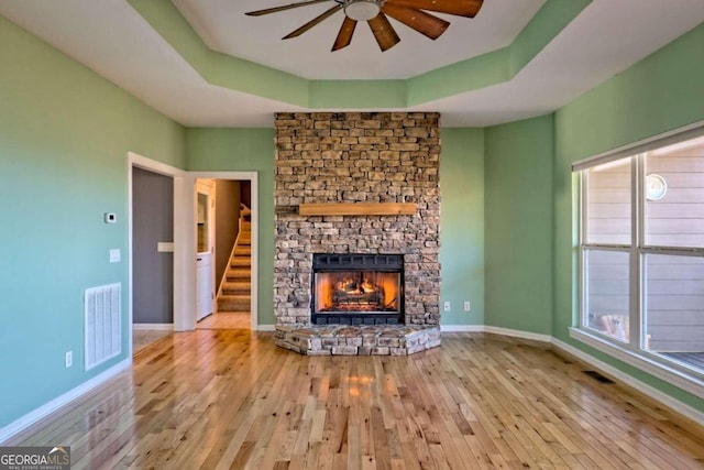 unfurnished living room with a stone fireplace, ceiling fan, light hardwood / wood-style floors, and a raised ceiling