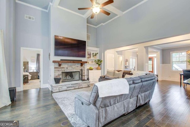 living room featuring a high ceiling, ceiling fan, a stone fireplace, crown molding, and dark wood-type flooring