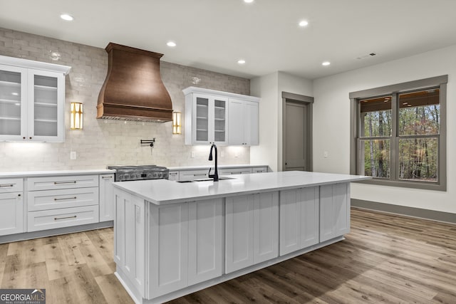 kitchen with sink, a kitchen island with sink, custom range hood, white cabinetry, and light wood-type flooring