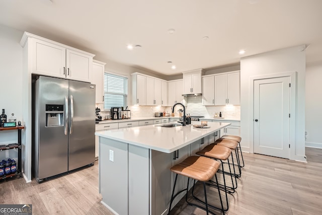 kitchen with stainless steel fridge, a center island with sink, white cabinets, light countertops, and a sink