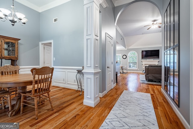 dining space featuring light wood-type flooring, decorative columns, ceiling fan with notable chandelier, crown molding, and a high ceiling