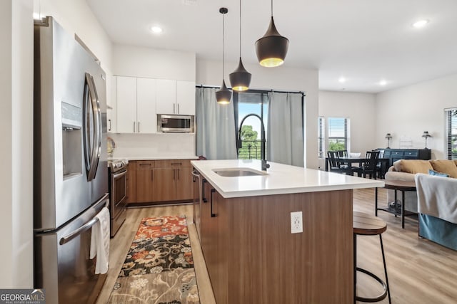 kitchen with white cabinetry, appliances with stainless steel finishes, light wood-type flooring, pendant lighting, and a kitchen island with sink