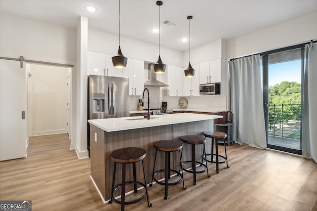 kitchen featuring a barn door, wall chimney range hood, white cabinetry, appliances with stainless steel finishes, and decorative light fixtures