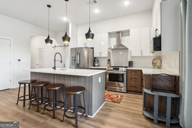 kitchen with a center island with sink, white cabinets, wall chimney range hood, light wood-type flooring, and appliances with stainless steel finishes