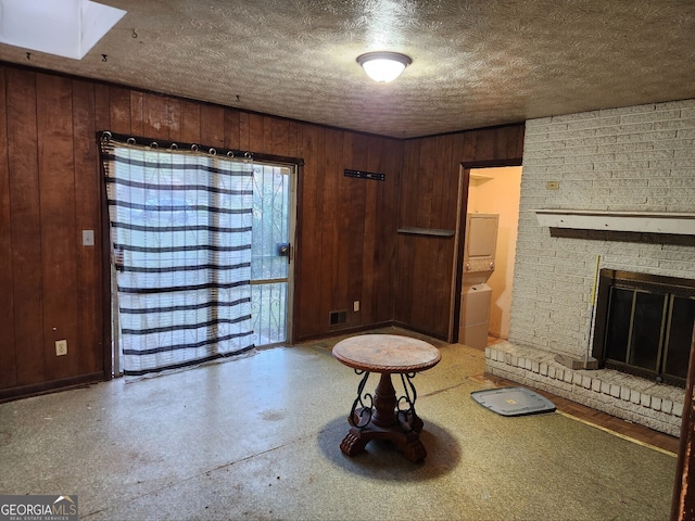 unfurnished living room with a textured ceiling, wooden walls, a brick fireplace, stacked washer / drying machine, and a skylight