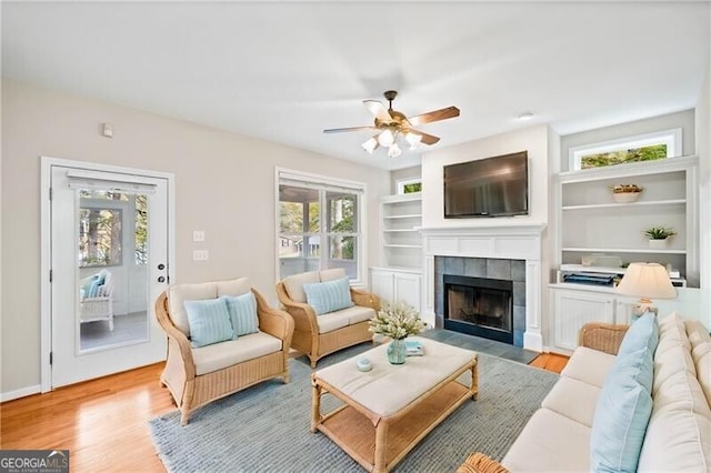 living room with a tiled fireplace, light hardwood / wood-style flooring, ceiling fan, and built in features