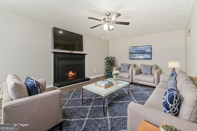 living room with a brick fireplace, dark wood-type flooring, a textured ceiling, and ceiling fan