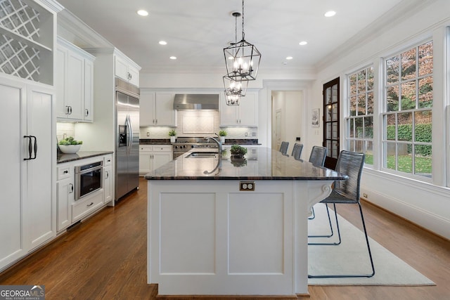 kitchen with white cabinetry, wall chimney exhaust hood, built in fridge, an island with sink, and a breakfast bar area