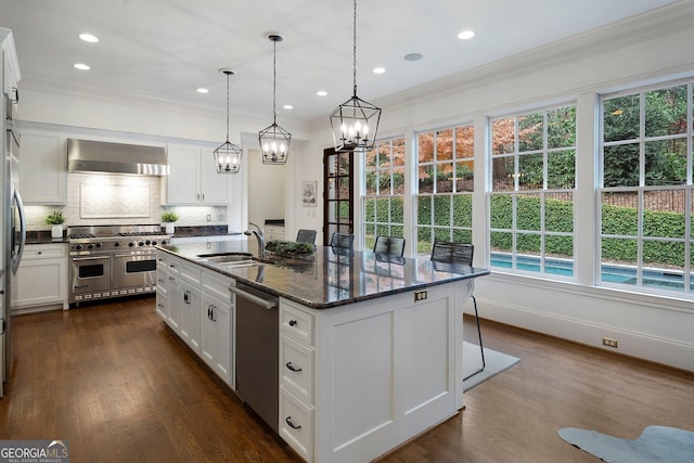 kitchen with wall chimney exhaust hood, pendant lighting, a kitchen island with sink, white cabinets, and appliances with stainless steel finishes