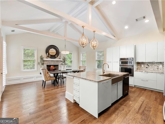 kitchen featuring a warm lit fireplace, stainless steel double oven, visible vents, light stone countertops, and an island with sink