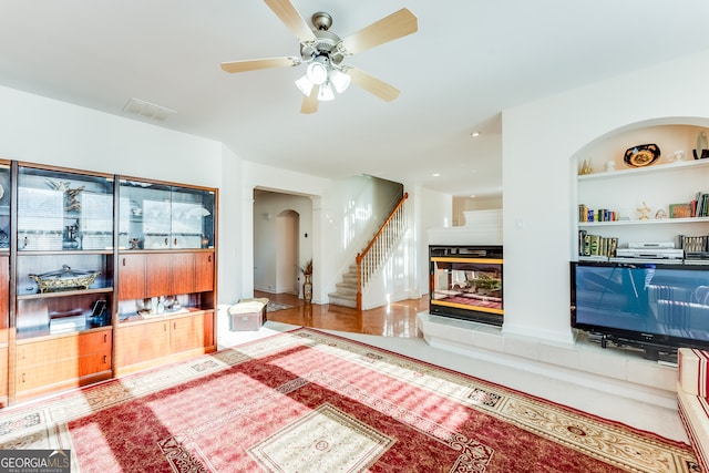 living room with ceiling fan, a tiled fireplace, and built in shelves