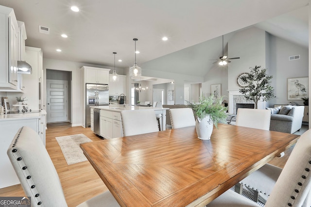 dining space featuring light hardwood / wood-style floors, sink, ceiling fan with notable chandelier, high vaulted ceiling, and a brick fireplace