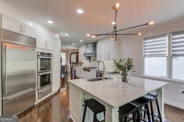 kitchen featuring a large island with sink, appliances with stainless steel finishes, wall chimney exhaust hood, decorative light fixtures, and white cabinets