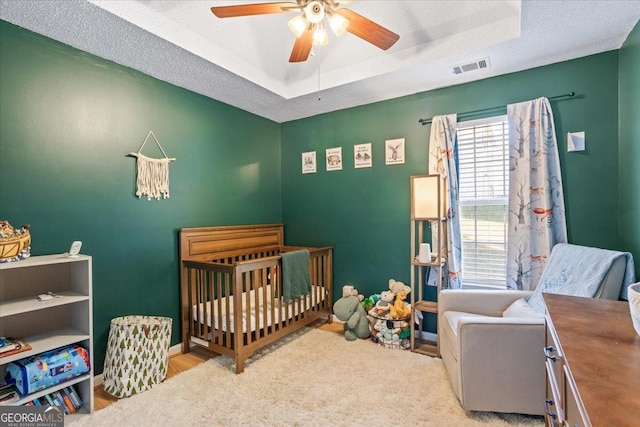 bedroom featuring ceiling fan, a textured ceiling, a tray ceiling, light hardwood / wood-style floors, and a crib