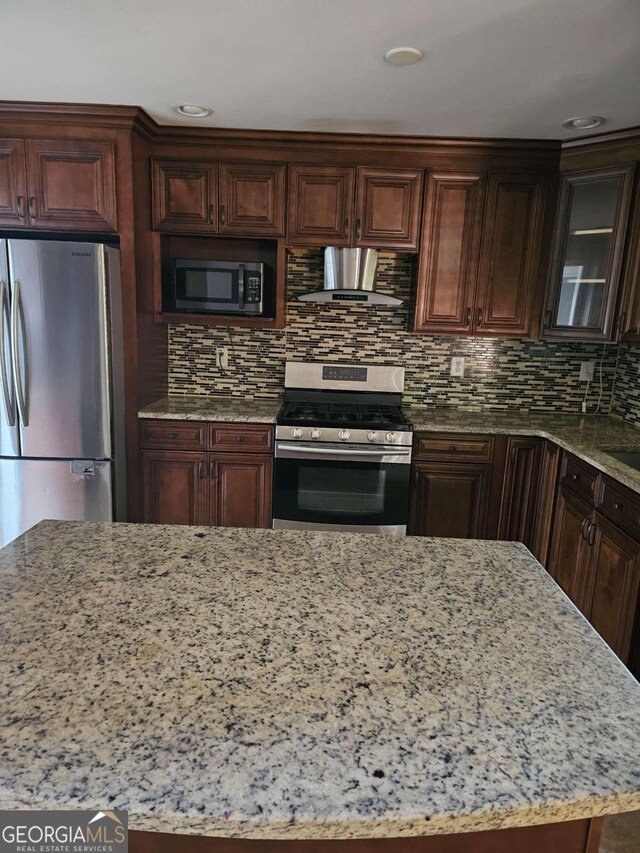 kitchen with light stone counters, dark brown cabinets, stainless steel appliances, and wall chimney range hood