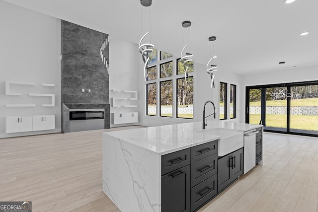 kitchen with sink, hanging light fixtures, light stone counters, an island with sink, and light wood-type flooring