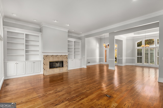 unfurnished living room with built in shelves, crown molding, a tile fireplace, hardwood / wood-style flooring, and an inviting chandelier