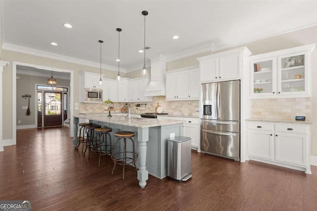 kitchen featuring a center island with sink, hanging light fixtures, appliances with stainless steel finishes, custom range hood, and white cabinets