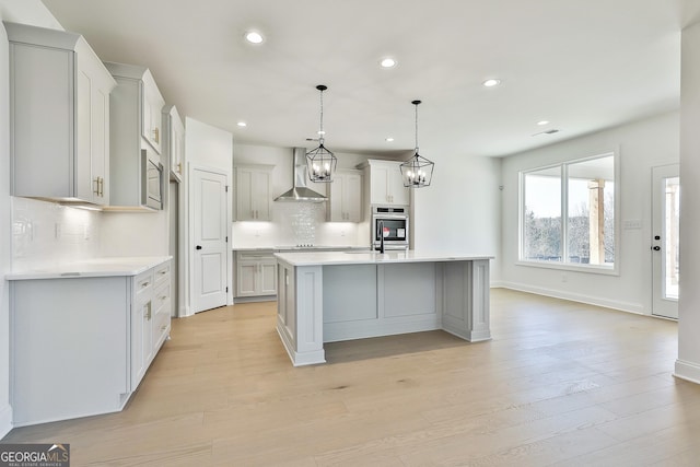 kitchen with wall chimney exhaust hood, stainless steel appliances, pendant lighting, light hardwood / wood-style floors, and a kitchen island with sink