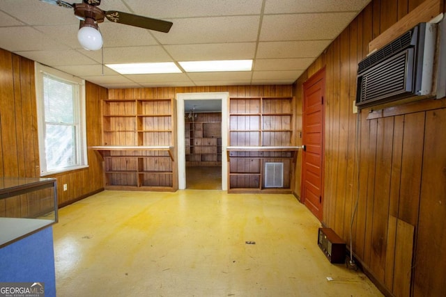 unfurnished living room with a drop ceiling, wood walls, ceiling fan, built in shelves, and a wall unit AC